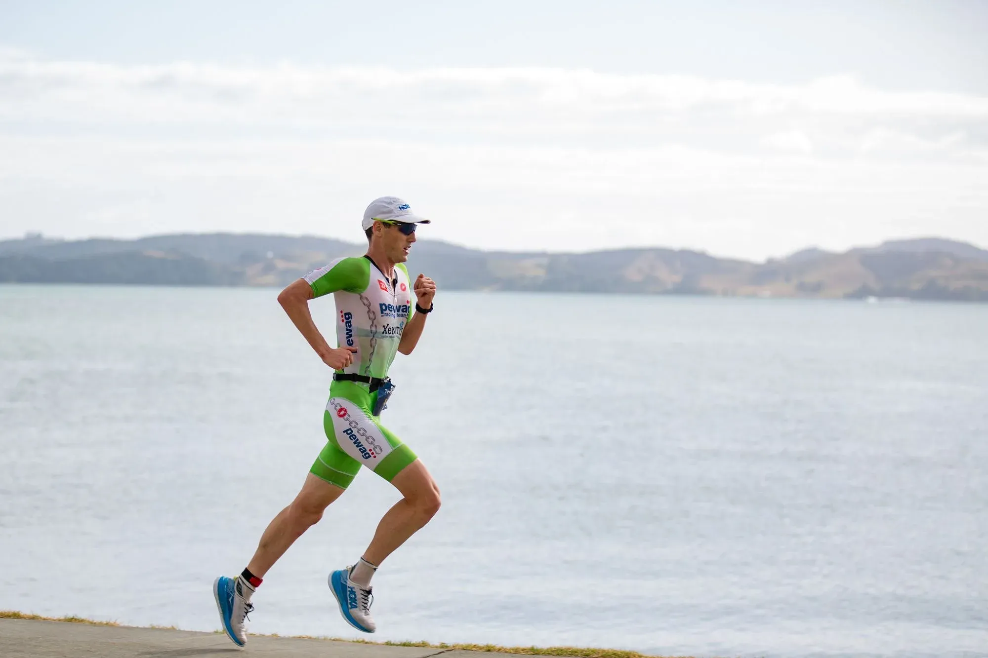 A triathlete in a green and white race suit running along the water's edge, hills behind